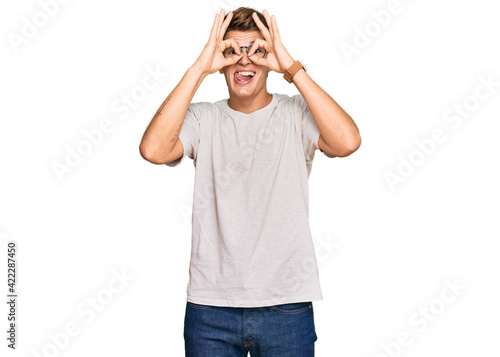 Handsome caucasian man wearing casual clothes and glasses doing ok gesture like binoculars sticking tongue out, eyes looking through fingers. crazy expression.