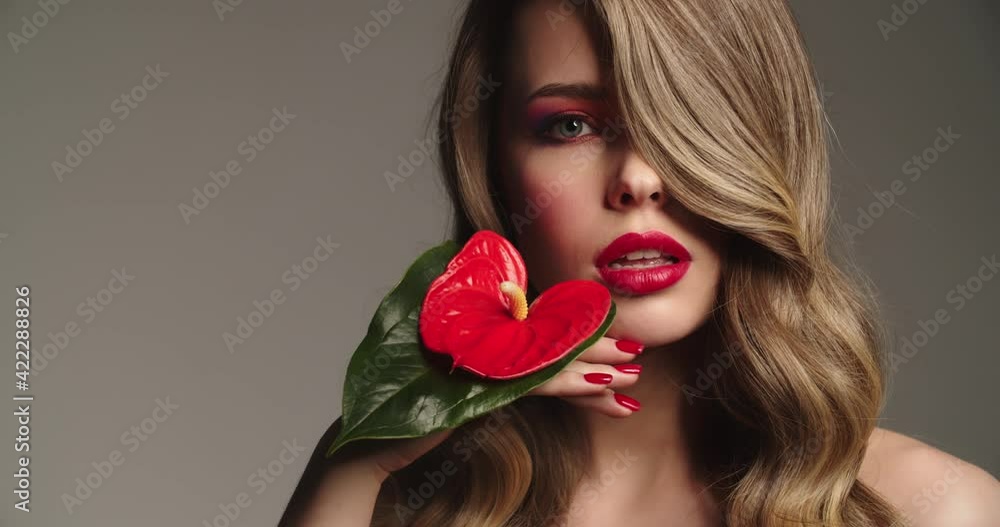 Sexy blonde woman with red lips holds a red flower. Fashion model with a long wavy hair posing at studio. Closeup face of a pretty young woman with bright makeup. Slow motion. Sexy blonde woman with red lips holds a red flower. Fashion model with a long wavy hair posing at studio. Closeup face of a pretty young woman with bright makeup. Slow motion.