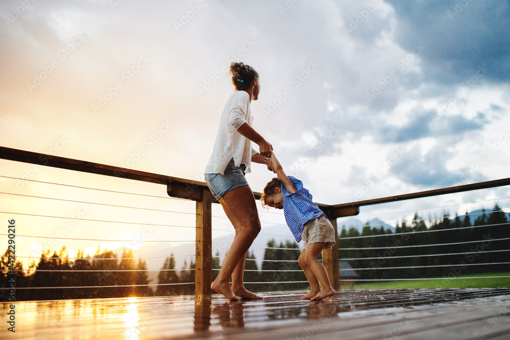 Mother with small daughter playing in rain on patio of wooden cabin, holiday in nature concept. - obrazy, fototapety, plakaty 