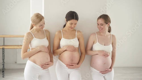 Three young pregnant women standing in studio in white sportive outfit with naked tummy, future moms stand talking, hugging their belly.