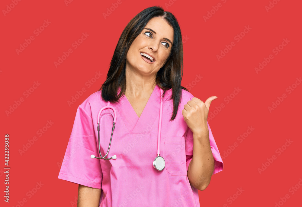 Middle age brunette woman wearing doctor uniform and stethoscope smiling with happy face looking and pointing to the side with thumb up. Middle age brunette woman wearing doctor uniform and stethoscope smiling with happy face looking and pointing to the side with thumb up.