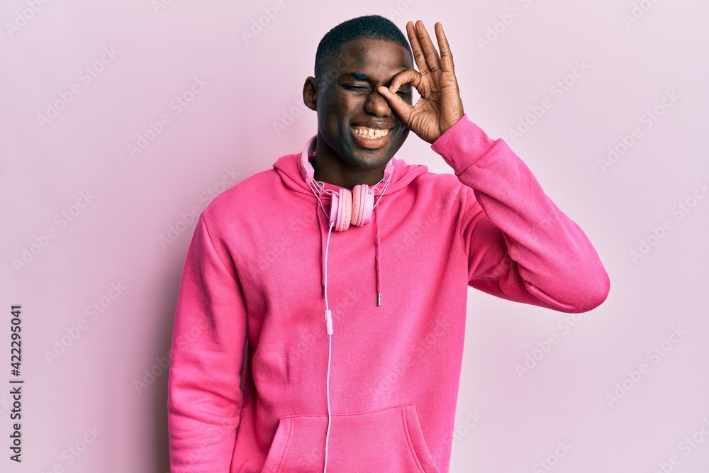 Young african american man wearing gym clothes and using headphones doing ok gesture with hand smiling, eye looking through fingers with happy face. Young african american man wearing gym clothes and using headphones doing ok gesture with hand smiling, eye looking through fingers with happy face.