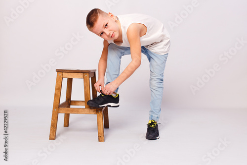 Nine-year-old European boy puts his foot on stool and ties his shoelaces on a white background