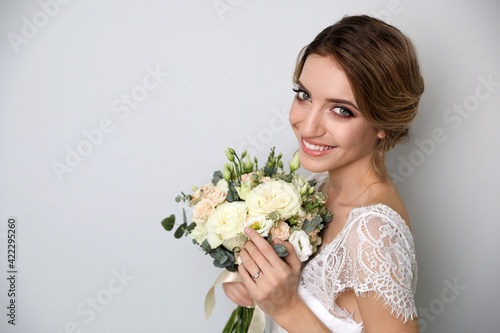 Young bride with elegant hairstyle holding wedding bouquet on light grey background. Space for text