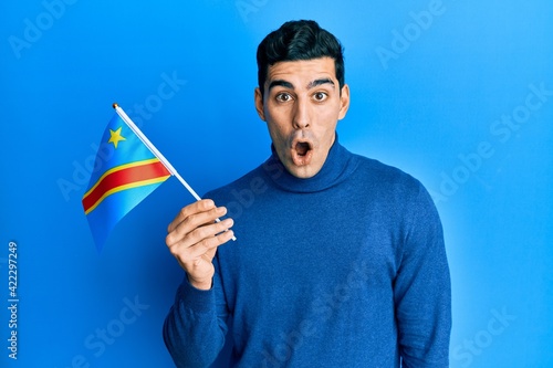 Handsome hispanic man holding democratic republic of the congo flag scared and amazed with open mouth for surprise, disbelief face