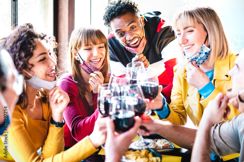 Multiracial friends toasting red wine at restaurant bar wearing open face mask - New normal life style concept with happy people having fun together after lockdown reopening - Bright vivid filter