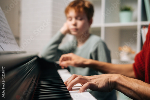 Focused redhead child student boy listening his teacher play the piano during lesson. Father teaching son to play musical instrument in living room. Concept of music education.