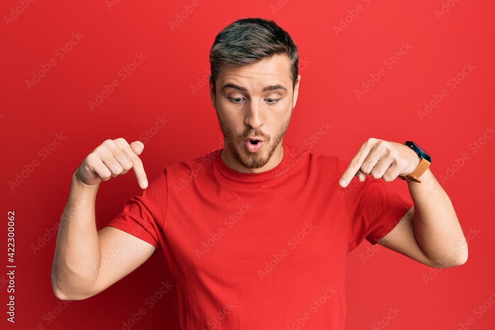 Handsome caucasian man wearing casual red tshirt pointing down with fingers showing advertisement, surprised face and open mouth Handsome caucasian man wearing casual red tshirt pointing down with fingers showing advertisement, surprised face and open mouth