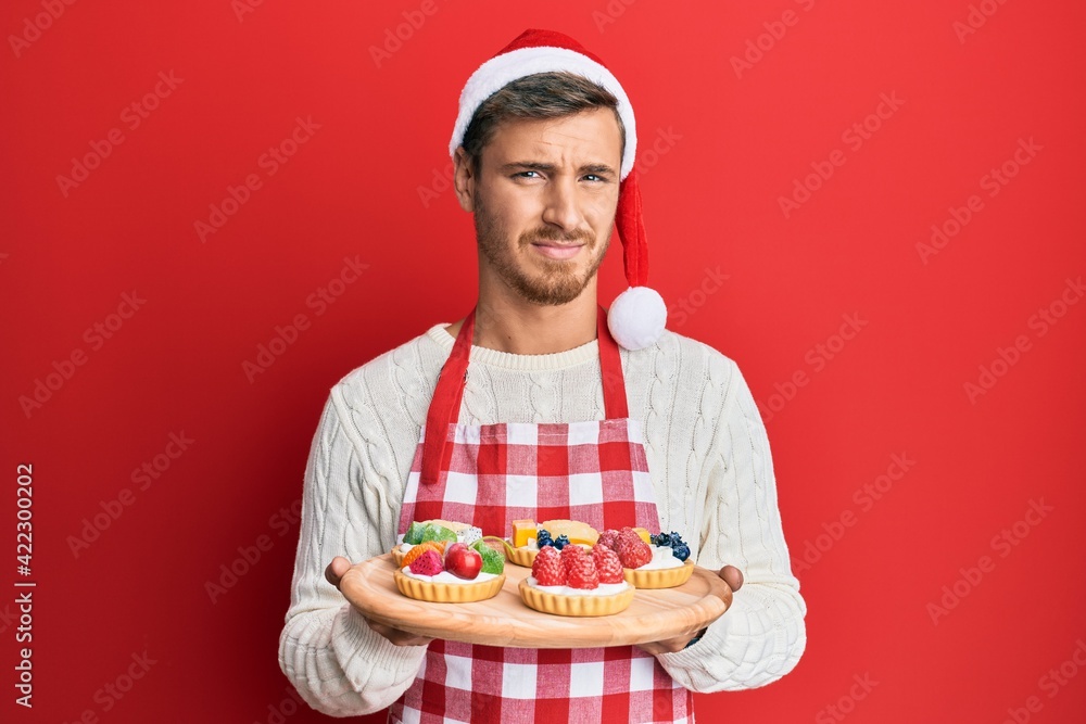 Handsome caucasian man wearing baker apron and christmas hat holding cakes clueless and confused expression. doubt concept. Handsome caucasian man wearing baker apron and christmas hat holding cakes clueless and confused expression. doubt concept.