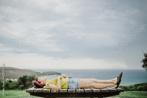 mujer joven recostada boca arriba sobre un banco de madera en un mirador con la playa de fondo con cielo nueblado photo