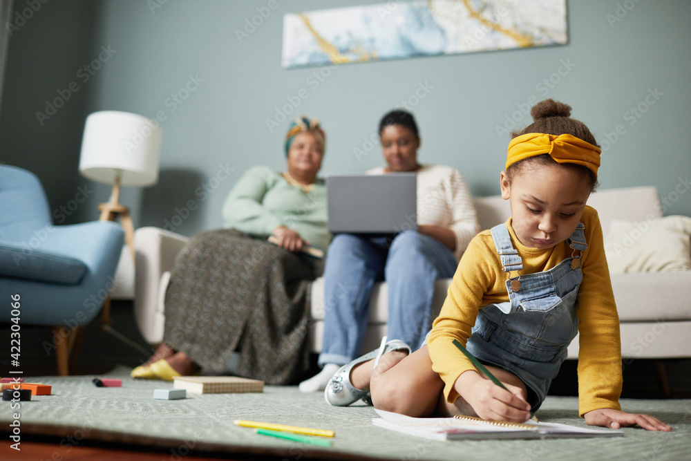 Front view portrait of cute African-American girl drawing while sitting on floor in home interior with family in background, copy space - obrazy, fototapety, plakaty 