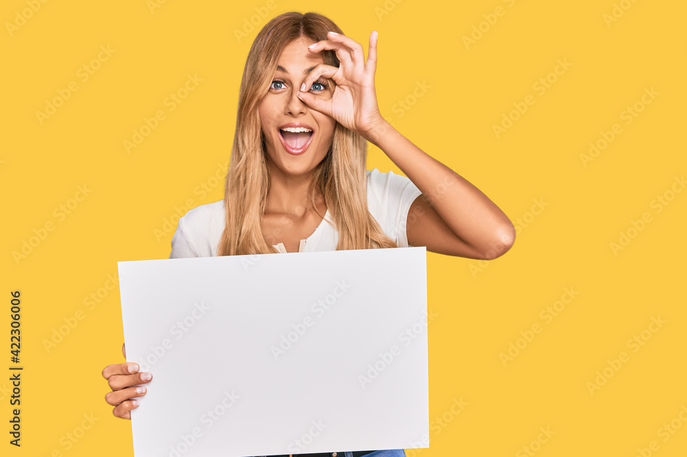Beautiful blonde young woman holding blank empty banner smiling happy doing ok sign with hand on eye looking through fingers Beautiful blonde young woman holding blank empty banner smiling happy doing ok sign with hand on eye looking through fingers