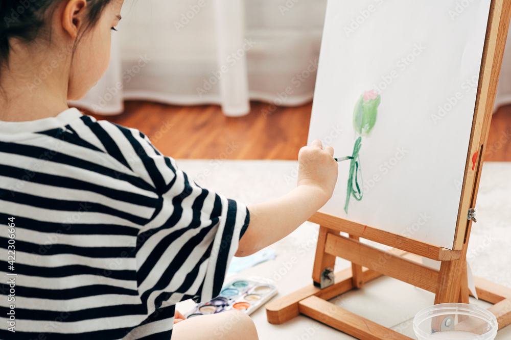 Rear view image of a little girl kid painting a flower on the easel at home. Cute kid sitting on the carpet and drawing in her room.
