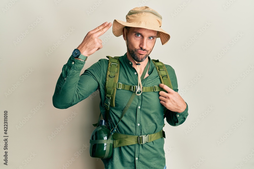 Fototapeta premium Handsome man with beard wearing explorer hat and backpack shooting and killing oneself pointing hand and fingers to head like gun, suicide gesture.