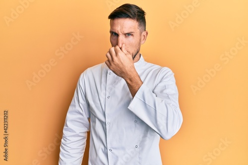 Handsome man with beard wearing professional cook uniform smelling something stinky and disgusting, intolerable smell, holding breath with fingers on nose. bad smell