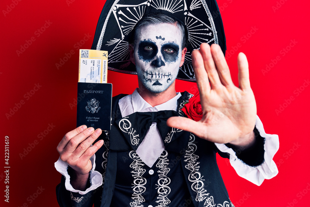 Young man wearing mexican day of the dead costume holding united states passport and boarding pass with open hand doing stop sign with serious and confident expression, defense gesture