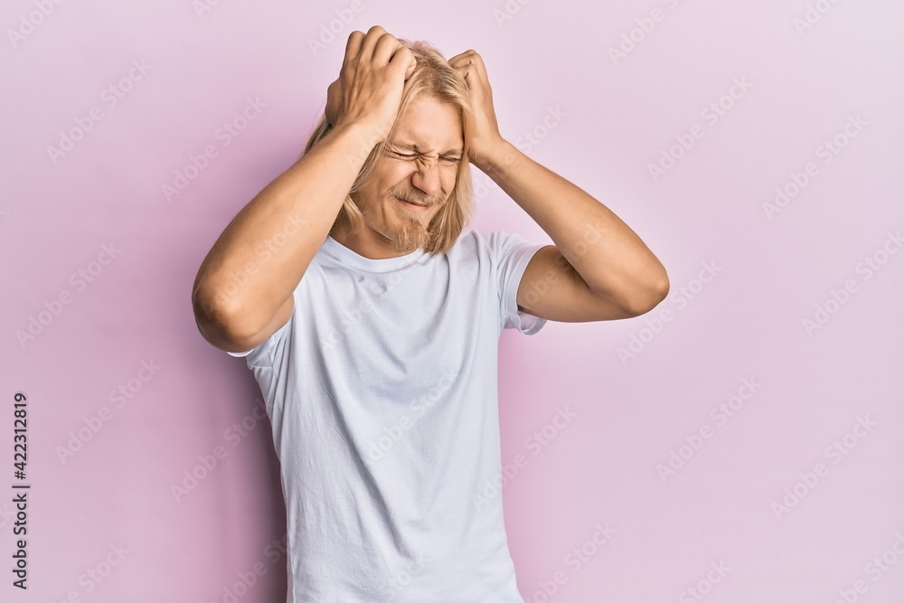 Caucasian young man with long hair wearing casual white t shirt suffering from headache desperate and stressed because pain and migraine. hands on head. Caucasian young man with long hair wearing casual white t shirt suffering from headache desperate and stressed because pain and migraine. hands on head.