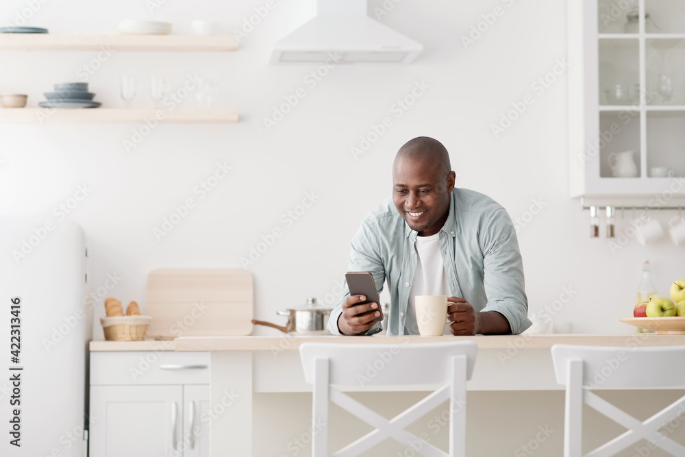 Happy african american man using smartphone and having morning coffee in the kitchen Happy african american man using smartphone and having morning coffee in the kitchen