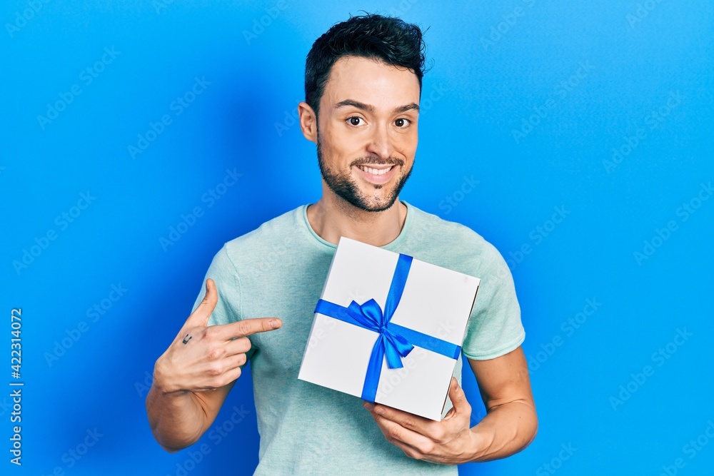 Young hispanic man holding gift smiling happy pointing with hand and finger Young hispanic man holding gift smiling happy pointing with hand and finger