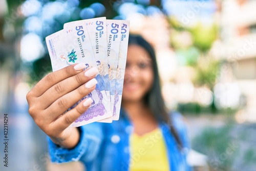 Young latin woman smiling happy holding colombian pesos at city.