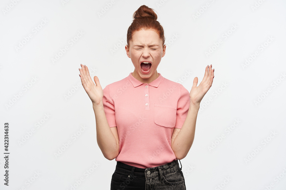 Angry looking woman, screaming redhead girl with hair bun. Wearing pink t-shirt and black jeans. Raising her hands and yell in anger with closed eyes. Stand isolated over white background Angry looking woman, screaming redhead girl with hair bun. Wearing pink t-shirt and black jeans. Raising her hands and yell in anger with closed eyes. Stand isolated over white background