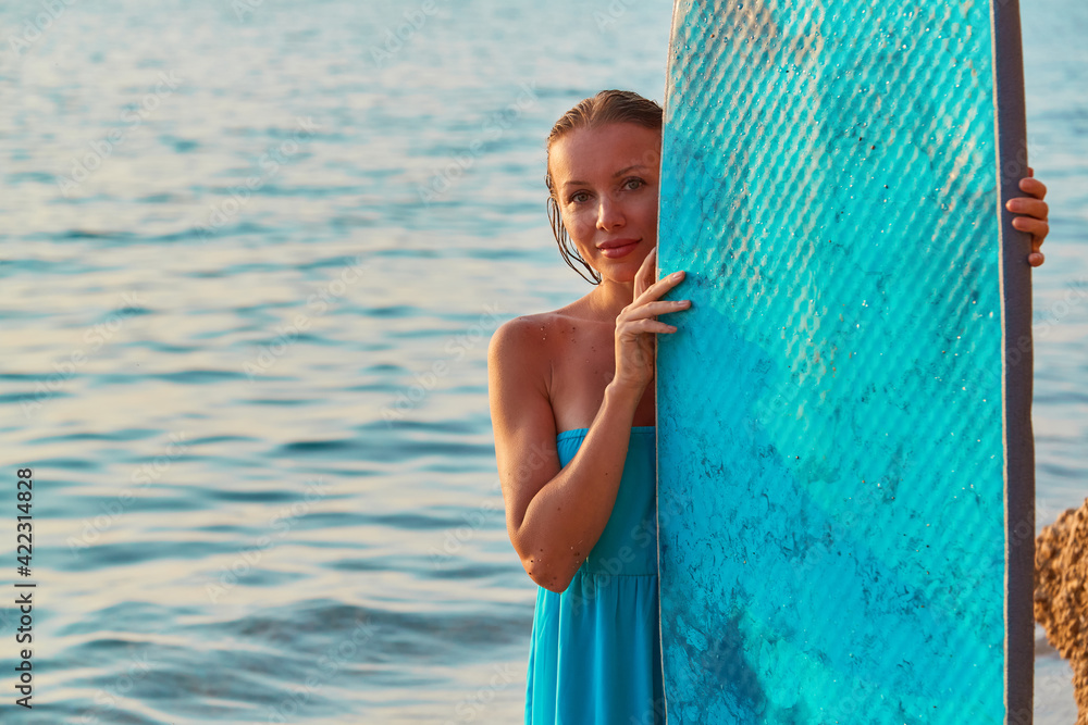 Smiling woman posing with surfboard an seashore. Happy girl surfer in blue dress. Summertime and time to adventure idea