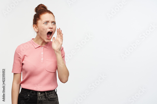 Portrait of shocked girl with ginger hair bun. Wearing pink t-shirt and black jeans. Shouts , holds palm next to mouth and watching to the right at copy space, isolated over white background
