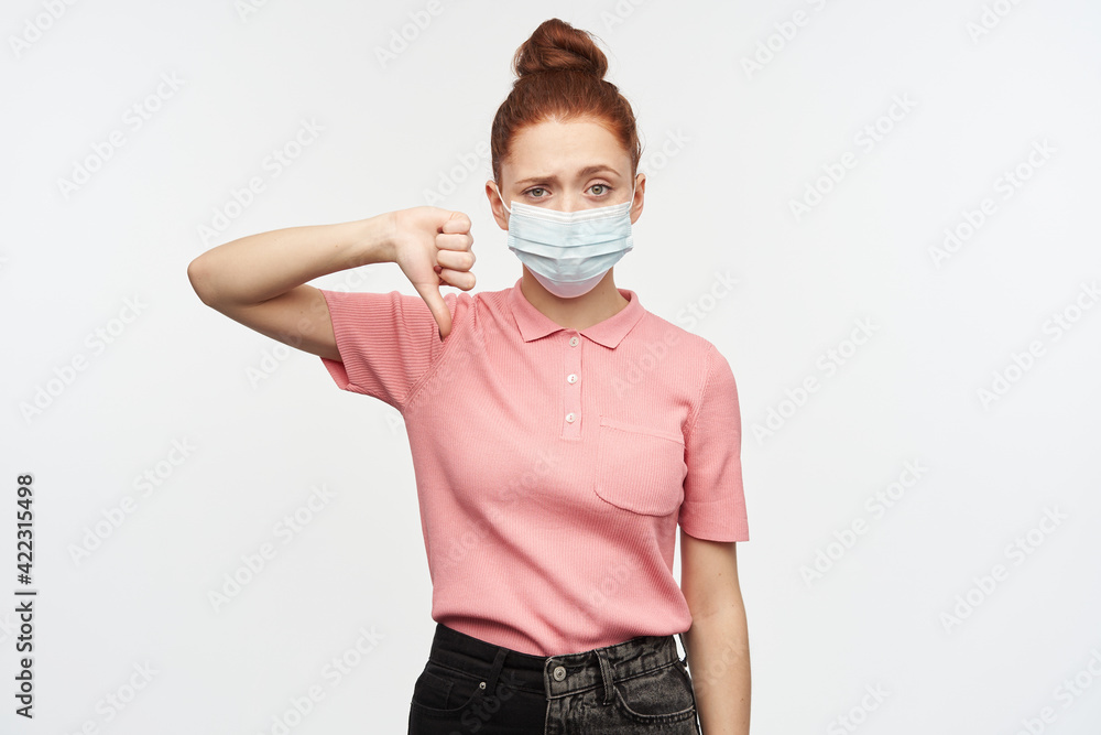 Portrait of negative, unhappy girl with ginger hair bun. Wearing pink t-shirt and medical, protective face mask. Shows thumb down sign. Watching at the camera, isolated over white background. Portrait of negative, unhappy girl with ginger hair bun. Wearing pink t-shirt and medical, protective face mask. Shows thumb down sign. Watching at the camera, isolated over white background.