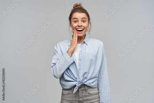 Lovely, beautiful blond girl with hair bun. Wearing blue striped knotted shirt. Emotion concept. Touching her cheek. Happy to hear compliment. Watching at the camera, isolated over grey background