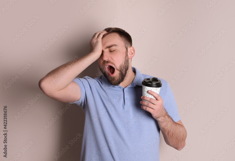 Sleepy young man with cup of coffee yawning on beige background Sleepy young man with cup of coffee yawning on beige background