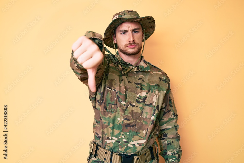 Young caucasian man wearing camouflage army uniform looking unhappy and angry showing rejection and negative with thumbs down gesture. bad expression. Young caucasian man wearing camouflage army uniform looking unhappy and angry showing rejection and negative with thumbs down gesture. bad expression.