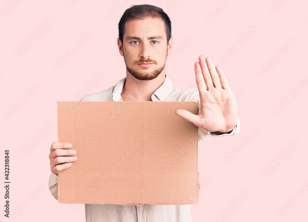 Young handsome caucasian man holding empty cardboard banner with open hand doing stop sign with serious and confident expression, defense gesture Young handsome caucasian man holding empty cardboard banner with open hand doing stop sign with serious and confident expression, defense gesture