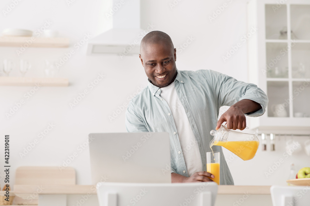 Mature black man having video chat with family on laptop, pouring juice in the kitchen