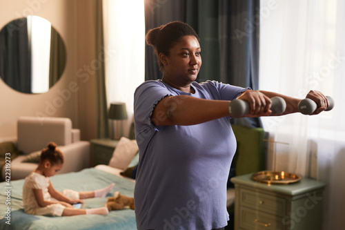 Waist up portrait of young African-American mother working out with dumbbells at home with little girl in background, copy space