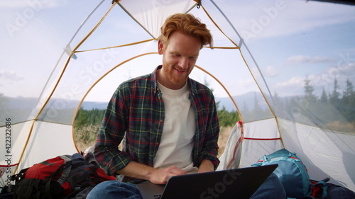 Redhead guy using laptop during hike. Male freelancer working on laptop in tent