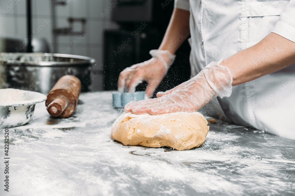 Female hands knead dough on the table next to a piece of dough and a baking dish. 