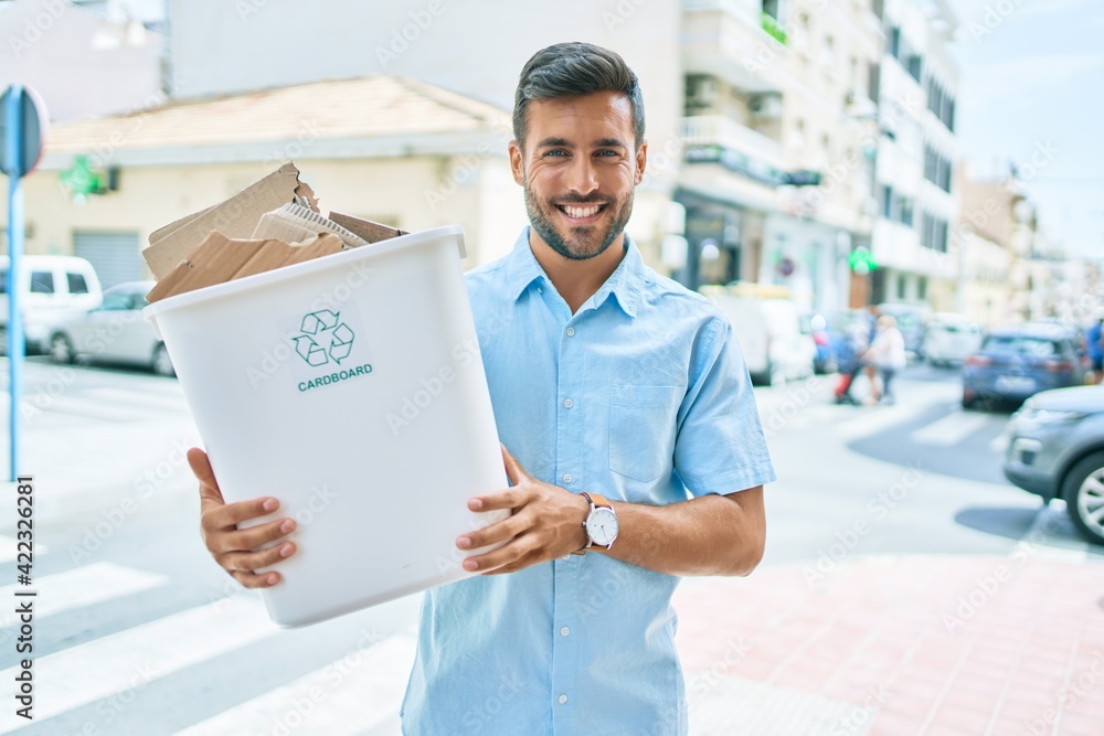 Young hispanic man smiling happy recycling. Holding full bin of cardboard at street of city.