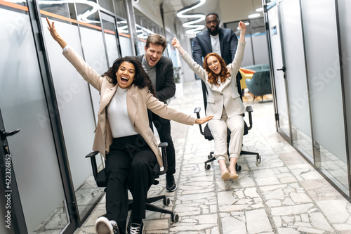 Joyful male and female colleagues take a break from work, having fun. Happy multiracial office employees in formal wear making a race on chairs, smiling and laughing