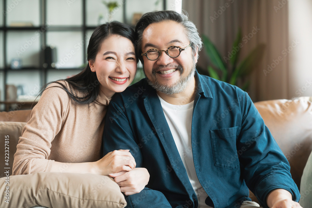 Portrait of Asian mature couple sitting and smiling in living room. wife woman hand hold husband arm from behind and look at camera with happiness and cheerful safty amd insurance family concept Portrait of Asian mature couple sitting and smiling in living room. wife woman hand hold husband arm from behind and look at camera with happiness and cheerful safty amd insurance family concept