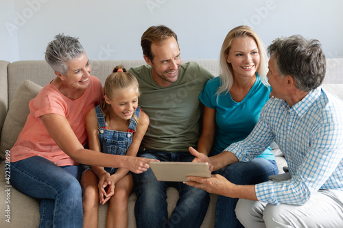 Smiling caucasian grandparents on couch with granddaughter and her parents looking at tablet