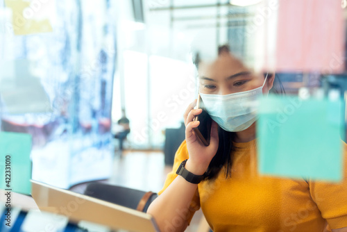 creative agency asian female casual cloth and face mask protection conversation with smartphone freelance working with laptop at coworking area office space with freshness with blur office background