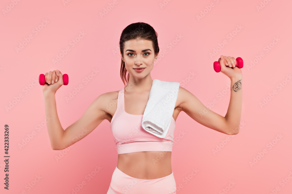 Young sportswoman with towel exercising with dumbbells isolated on pink Young sportswoman with towel exercising with dumbbells isolated on pink