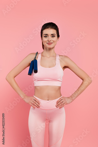 Smiling sportswoman with jump rope and hands on hips isolated on pink