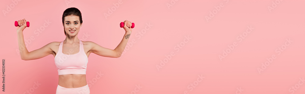 Brunette sportswoman exercising with dumbbells and smiling at camera isolated on pink, banner Brunette sportswoman exercising with dumbbells and smiling at camera isolated on pink, banner