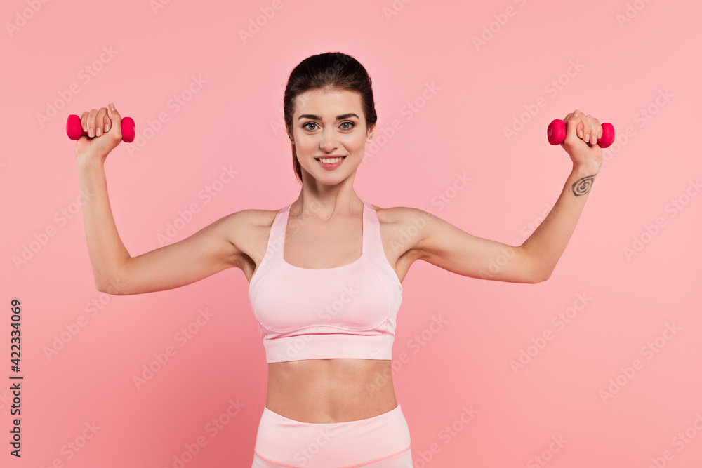 Cheerful sportswoman training with dumbbells isolated on pink Cheerful sportswoman training with dumbbells isolated on pink