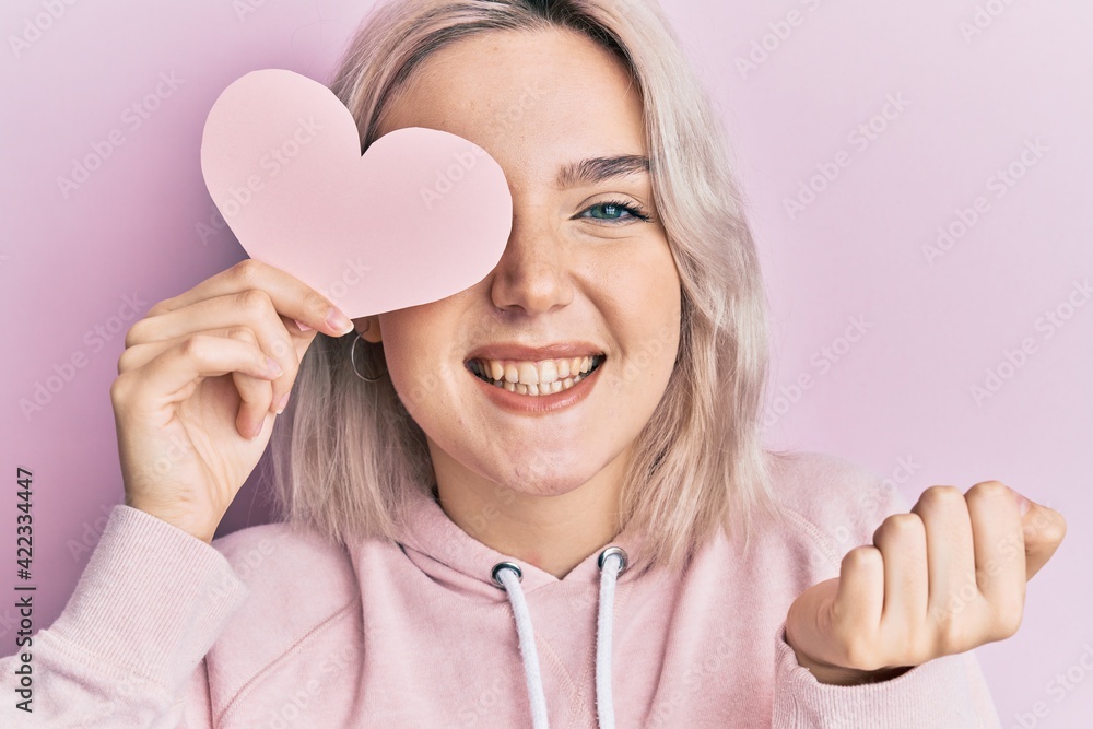 Young blonde girl holding heart shape paper over eye screaming proud, celebrating victory and success very excited with raised arm Young blonde girl holding heart shape paper over eye screaming proud, celebrating victory and success very excited with raised arm