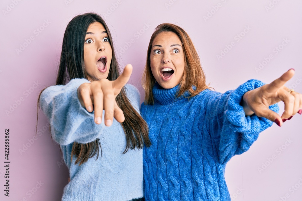 Hispanic family of mother and daughter wearing wool winter sweater pointing with finger surprised ahead, open mouth amazed expression, something on the front Hispanic family of mother and daughter wearing wool winter sweater pointing with finger surprised ahead, open mouth amazed expression, something on the front