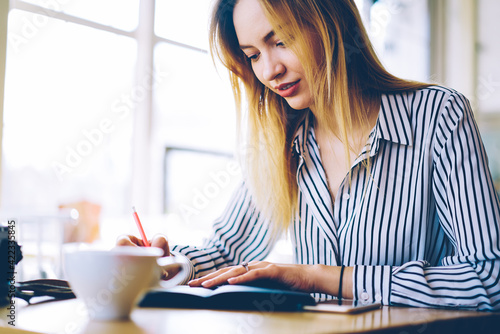 Intelligent Caucasian student in smart casual clothing writing memo notes in education textbook, clever female 20s creating text while doing university homework using notepad in cafe interior