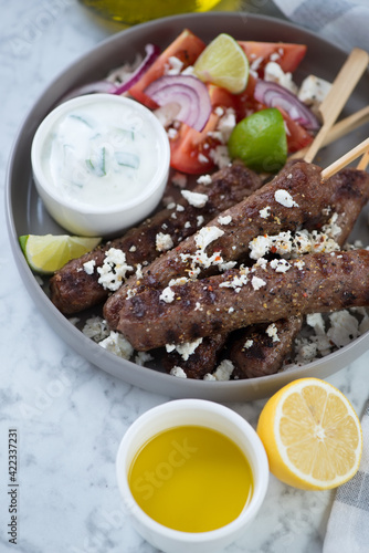 Close-up of bbq greek kofta kabobs with tzatziki sauce, feta and vegetables, vertical shot on a grey marble surface