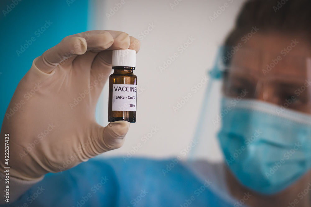 doctor in a protective shield, veil, and sterile gloved suit holds a single weapon against the Coronavirus pandemic. A scientist holds an ampoule against a Covid-19 and points to the camera.